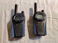 Two Cobra MicroTALK walkie talkies with circular screen and blue sides on a white cloth background