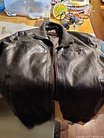 Color image of the Guess men's leather jacket laid out flat on a surface, showing the front with zipped pockets and rib-knit collar.