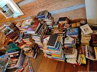 Photo showing multiple stacks of used books of varying sizes and genres arranged on a wooden floor against a wall