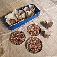 Photo showing six small cups and three small plates with intricate red, brown, gold floral designs on white ceramic. Cups are arranged in blue box with paper dividers.