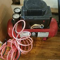 Front view of Jobmate air compressor showing motor housing, pressure gauges, and coiled pink air hose with black power cord on wooden floor.