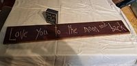 Photo of large wall hanging with phrase 'Love you to the moon and back' in white script on dark wood and small matching bookshelf piece near it.