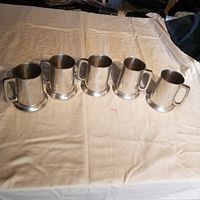 Five used pewter mugs arranged side by side on a beige fabric surface, showing their shape and handles.