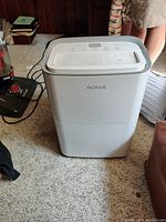White NOMA portable air conditioner, showing top control panel and vents, set on carpeted floor near some household items.