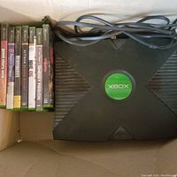 Photo showing the Original Xbox console and six Xbox game cases stacked vertically alongside the console inside a cardboard box. The power cable is coiled and placed on top of the console.