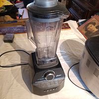 Oster Versa blender on a table with power cord visible, showing blender pitcher, lid, and base with controls.