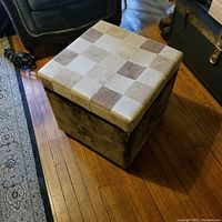Square footstool with checkered patterned lid and dark brown sides, sitting on wooden floor near furniture.
