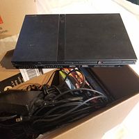 Photo of black Sony PlayStation 2 Slim console sitting over a box containing power cables, controllers, and game cases.