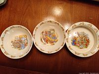 Photo of three ceramic Bunnykins bowls with detailed bunny scenes and decorative border