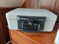 Photo of a gray portable security box with black handle and key lock, labeled Sentry Safe 1100.
