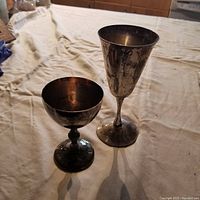 Two silver goblets, one shorter with a round bowl and decorative stem, the other taller with narrow bowl and straight stem, showing tarnish and wear.