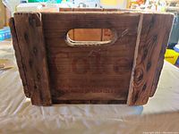 Photo of one side of the worn wooden crate showing the cutout handle and faded Coca-Cola logo in red lettering.
