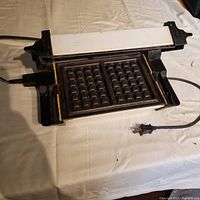 Vintage black and white rectangular Tefal waffle iron with grid pattern non-stick cooking plate and attached power cord visible on a cloth surface.