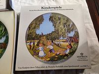 Close-up of one plate showing children playing in a forest clearing with hay and a swing, with detail text in German referencing a charity donation from purchase price and Heinrich brand logo, confirming plate's authenticity.