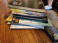 Stack of assorted vinyl records on wooden floor showing various album covers and sleeves, indicating a broad collection of used vinyl records.