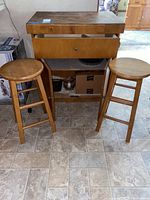 Photo shows wooden buffet with drawer partially open accompanied by two wooden stools on tile floor.