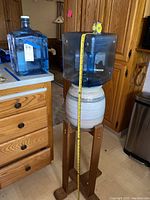 Photo showing clay water dispenser on wooden stand with blue square water jug on top and separate blue jug on countertop.