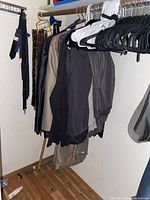 Full view of men's clothing on hangers in a closet, multiple black plastic and wooden hangers visible. Clothes include jackets and pants in various dark shades and beige.
