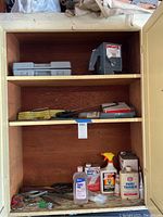 Full view inside the wooden cabinet showing shelves filled with various hand tools, measuring devices, and cleaning supplies including bottles of paint thinner, baby oil, and water seal.
