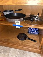 Cabinet shelf showing multiple pieces of cookware including frying pans, a small pan, and a glass lid.