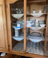 Wooden cabinet with glass doors displaying several shelves of assorted dishware including glasses, cups, saucers, plates, and bowls. Shelves lined with lace doilies.
