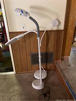 Two white adjustable floor lamps standing side by side against wood panel wall, with visible cords and bases. Lamps show curved flexible necks and modern design.