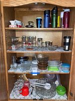 Wide shot of wooden shelf displaying various glassware and drinkware including goblets, small bowls, ceramic creamers, pitchers, and plastic travel mugs.