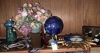 View of mixed home decor items on wooden surface including a flower arrangement in a green ceramic pot, dark blue globe on wooden stand, and partially visible sunburst wall clock