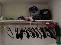 Closet shelf with multiple baseball caps stacked together alongside folded items and a set of various hangers hanging below on a rod.