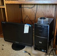Photo of Samsung monitor and HP desktop tower computer showing front and side, including cables and power connection, sitting on carpet with wooden desk background.