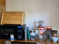 Photo showing the Sunbeam black microwave, wooden bread box on top, salt and pepper mills, and other kitchen items on a shelf against a beige patterned wall.