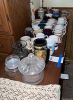 Photo showing a variety of coffee mugs and jars arranged on a table, including clear glass containers with lids and several colorful mugs.