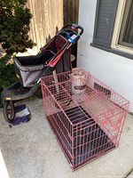 Photo showing Virago pet stroller with black and burgundy fabric and a pink metal pet cage with black base tray, both placed outdoors near a wall.