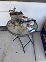 Full view of the round glass-top metal plant table with the planter, turtle figurines, mushroom garden ornament, decorative painted stones, and window chime laid out on top.
