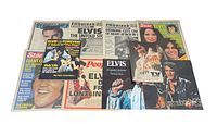 Photo showing a collection of Elvis Presley newspapers and magazines spread out, featuring covers with Elvis music and TV previews, multiple headlines and images.