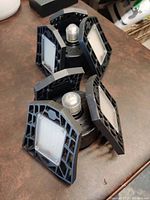 Two black LED lamps with three adjustable LED panels each, arranged flat for viewing. Each lamp has a central screw-in base and rectangular LED panel lights.