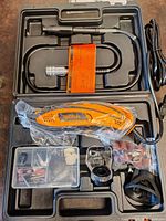 Opened plastic tool case showing orange and black electric rotary tool wrapped in plastic, a flexible shaft attachment, plastic accessory cases containing sanding discs, grinding stones, and polishing pads, a transparent protective guard, sanding bands in plastic bag, and a black keyless chuck.