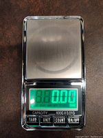 Digital pocket scale showing green backlit screen reading 0.00 with four buttons beneath and stainless steel weighing surface.