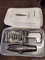 Complete view of the manicure and pedicure kit showing the handheld nail drill device, multiple drill bits and sanding bands arranged in plastic trays, power adapter, and carrying case with instruction manual.