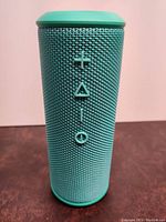 Front view showing control buttons for volume, play/pause, and power on teal mesh fabric Bluetooth speaker.