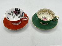 Two teacup and saucer sets together, showing the orange set with black and red rose design and the green set with pink rose and gold detailing.