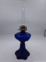 Full view of antique oil lamp in cobalt blue glass with clear chimney on neutral background.