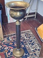 Full view of brass pot mounted on a tall pedestal placed on a patterned rug, showing overall shape and form