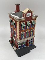 Angled front left view of the miniature post office building showing the brick pattern, windows, snow accents, wreath, presents, and snowman figure.