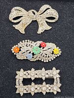 Photo showing three brooches on black background: bow-shaped brooch with clear rhinestones, oval filigree brooch with colorful flower accents, and rectangular rhinestone floral brooch.