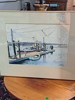 Framed watercolor painting depicting harbor dock, pilot boat, rowboat, and seagulls reflecting in water.