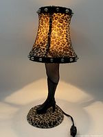 Full view of small decorative leg lamp illuminated, showing leopard print lampshade and black boot base with fishnet detail.