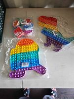 Two jumbo pop its, one dinosaur-shaped and one Among Us-shaped, rainbow-colored, in plastic packaging on a white table.