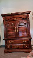 Front view of the wood armoire showing the cupboard with two doors and three drawers below with gold tone drawer pulls.