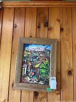 Framed original acrylic painting on canvas showing a colorful village scene hung on a wood-paneled wall.